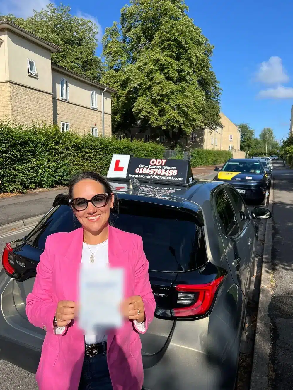 Smiling learner driver in a pink jacket holding her driving test pass certificate beside an Oxon Driving Tuitions automatic Toyota Yaris
