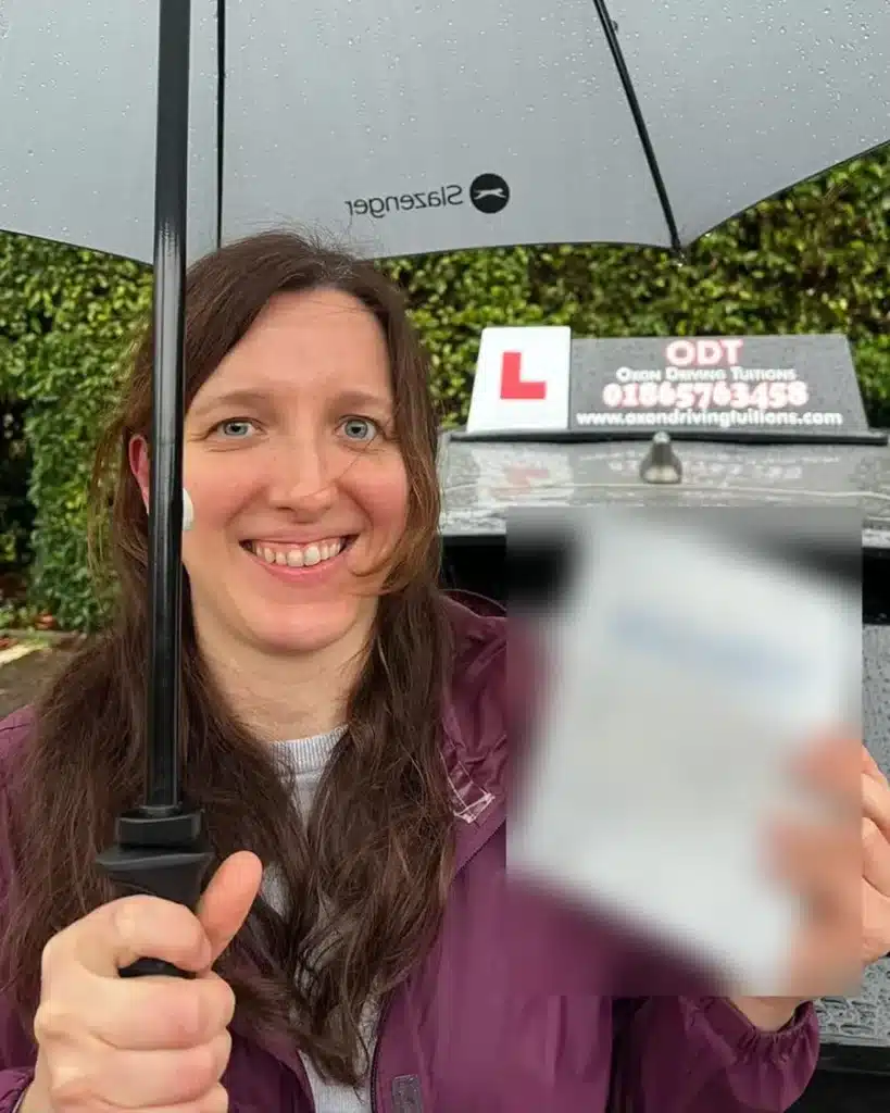 Radley learner driver smiling under an umbrella with her driving test pass certificate beside an Oxon Driving Tuitions car