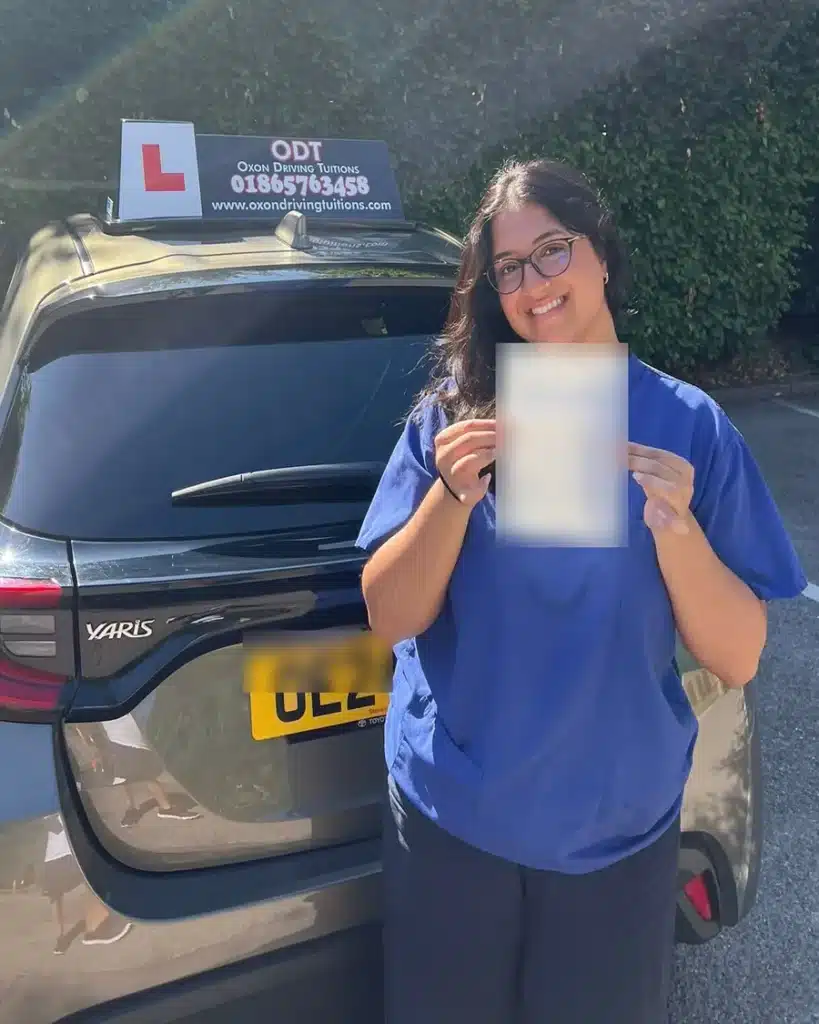 Doctor in scrubs smiling with her driving test pass certificate beside an Oxon Driving Tuitions automatic Toyota Yaris